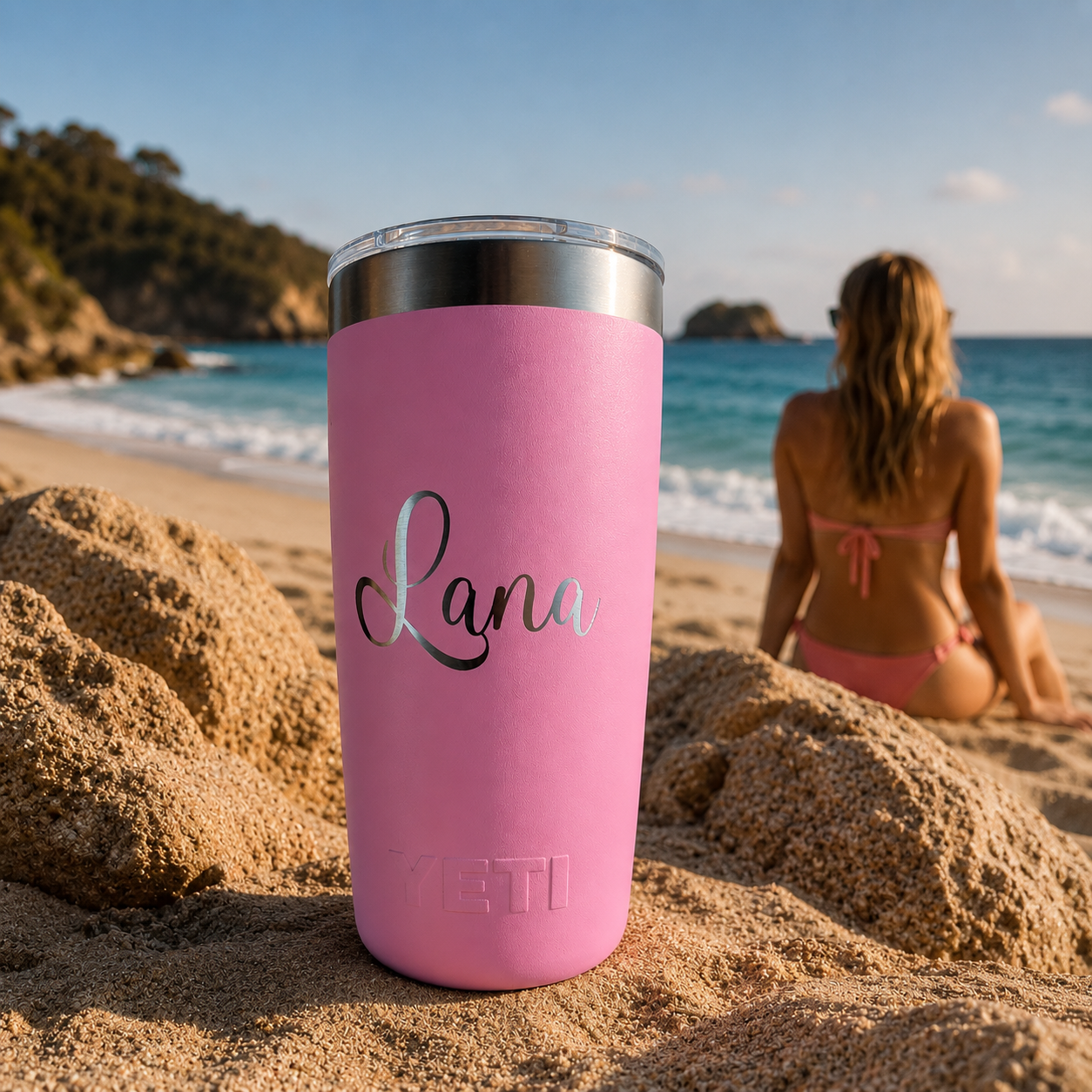 A pink insulated tumbler with a clear lid sits on sandy beach ground, with a woman in a pink bikini seated in the ocean chair area in the background. The tumbler has the name 'Lana' printed on it in silver script and 'YETI' branding near the bottom.